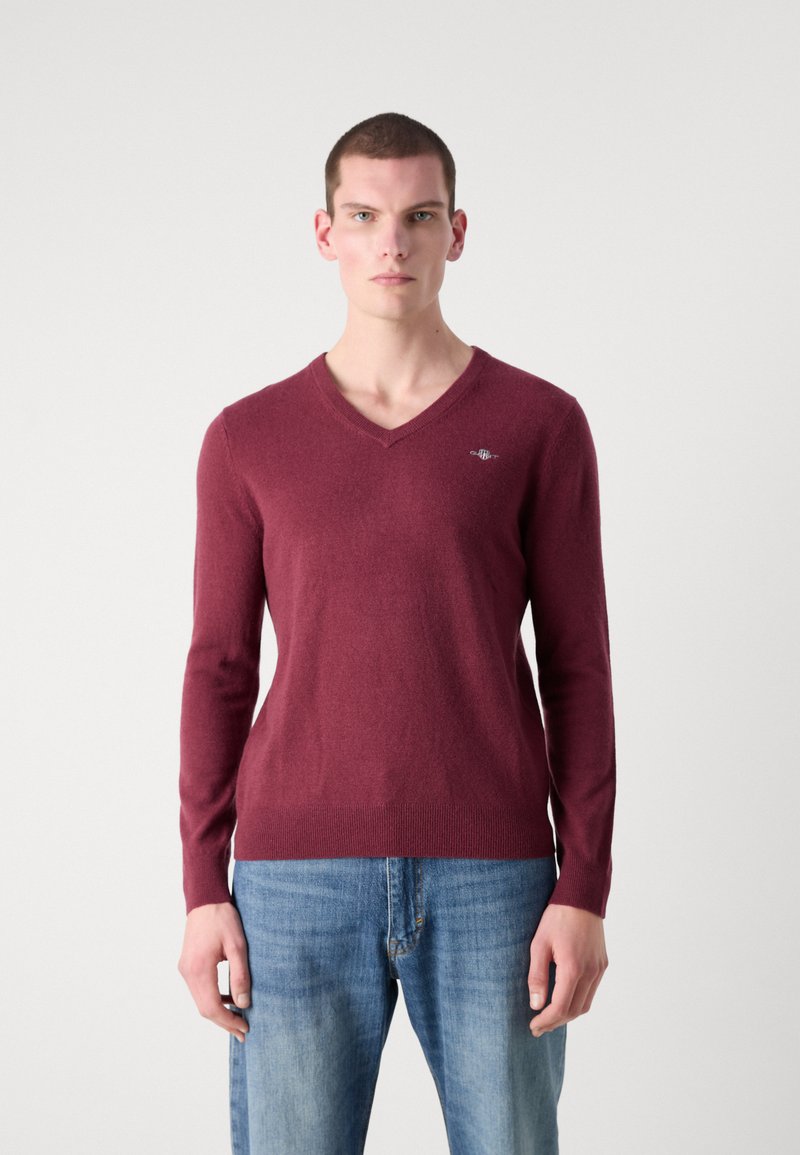 Suéter burdeos de cuello en V con mangas largas, hecho de una tela suave. Presenta un pequeño logotipo en el pecho y un diseño ajustado en la cintura.