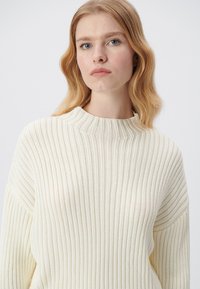 Cremfarbener Rippstrickpullover mit hohem Kragen und lockerer Passform. Hergestellt aus leichtem Strickmaterial mit vertikalen Streifen.