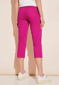Różowe spodnie capri o dopasowanym kroju i długości do kostek, z dwoma tylnymi kieszeniami i przeszyciami. Noszone z białymi sneakersami.