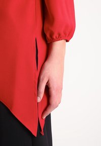 Blusa roja con mangas largas y abullonadas, que presenta una abertura en el costado y un dobladillo curvado. Hecha de un tejido ligero con textura suave.