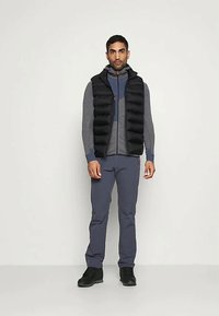 Mies, joka on pukeutunut mustaan untuvaliiviin harmaan vetoketjuhupparin päällä, tummansinsisiin housuihin ja mustiin saappaisiin, seisoo vaaleaa taustaa vasten.