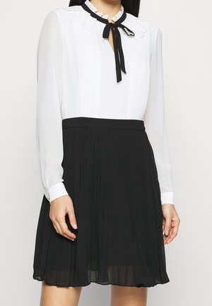 Frau trägt eine weiße langärmelige Bluse mit schwarzer Fliegenkragen und einem schwarzen plissierten Rock, steht vor einem schlichten Hintergrund.