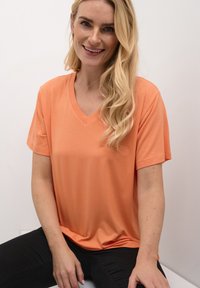 Short-sleeve, v-neck t-shirt in pastel orange fabric. Soft texture with a relaxed fit, paired with black pants. Simple design with no patterns.