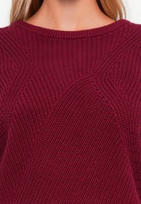 Pull en maille bordeaux avec des motifs texturés, doté d'un col rond et d'une finition lisse. Des coutures détaillées ajoutent un intérêt visuel.