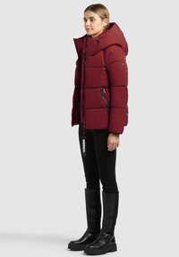 Femme portant une doudoune rouge à capuche, des leggings noirs et des bottes noires mi-mollet, debout de profil sur un fond gris uni.