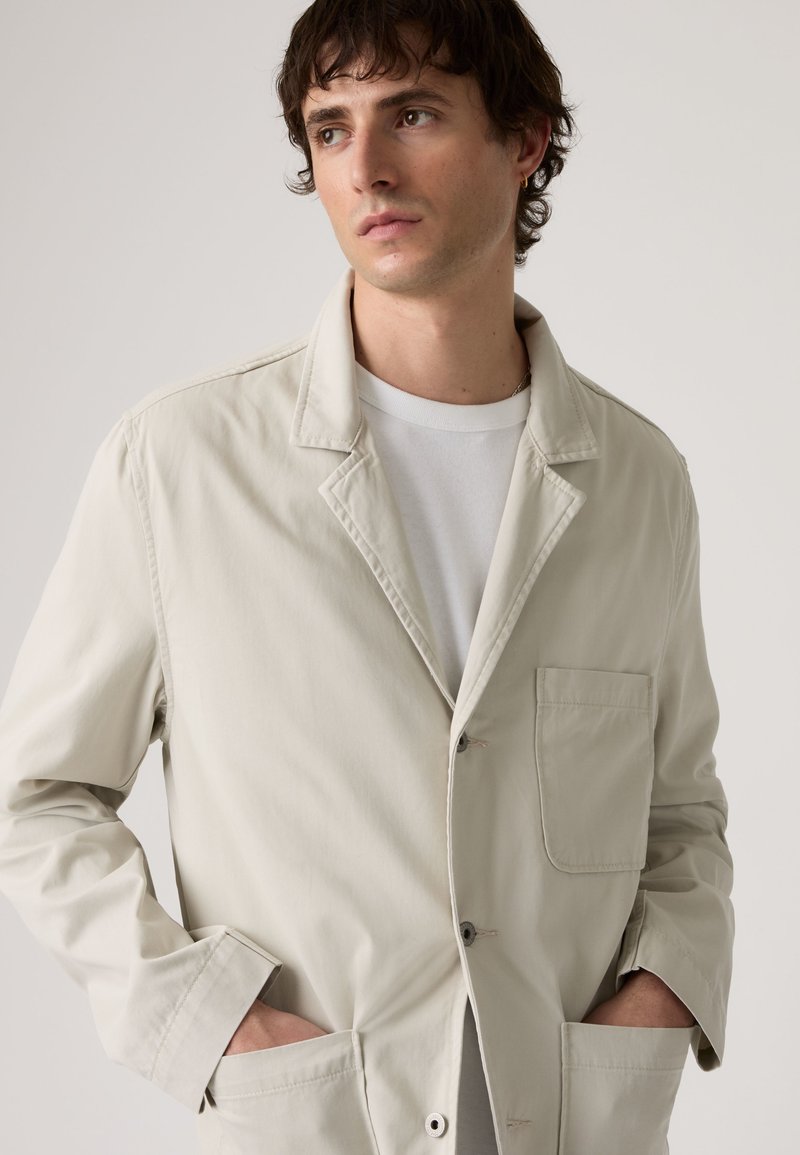 Jeune homme portant une veste beige clair sur un t-shirt blanc, les mains dans les poches, regardant légèrement sur le côté sur un fond uni.