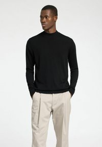 Schwarzer, langärmeliger, gerippter Strickpullover mit hohem Kragen, kombiniert mit hellbeigen, gefalteten Hosen. Glatte Textur, figurbetontes Design.
