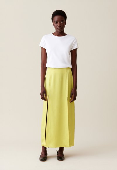 Camicia bianca a maniche corte abbinata a una lunga gonna gialla con fessure frontali. Le scarpe flatform nere completano l'outfit. Colori solidi, design minimale.