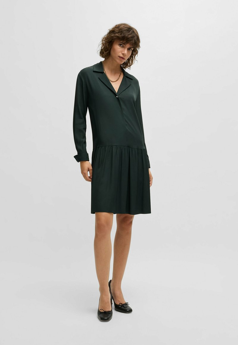Dunkelgrünes, langärmeliges Kleid aus einem glatten, satinähnlichen Stoff. Verfügt über einen Kragen, dezente Knopfdetails und eine geraffte Taille, das über dem Knie endet.