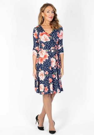 Frau trägt ein marineblaues Kleid mit weißen Polka-Dots und großen pinken Blumen, steht mit gekreuzten Beinen und schwarzen High Heels.
