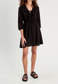 Robe noire avec un col à volants et des manches trois-quarts, taille froncée et jupe fluide ; assortie à des sandales noires à clous.