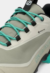 GORE-TEX tursko i lys grå og krem, med teal snørebånd, sorte detaljer og en teksturert overflate med en konturert form.