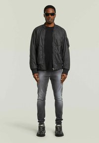 Giacca bomber nera con polsini a coste, abbinata a una camicia nera e jeans grigi sbiaditi. Il modello indossa occhiali da sole neri e sneakers nere dal design robusto.