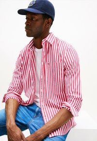 Camicia a righe rossa e bianca con maniche arrotolate, abbinata a una maglietta bianca e jeans in denim blu. La camicia ha una tasca sul petto.