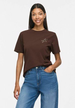 Kobieta z długimi ciemnymi włosami, ubrana w brązowy t-shirt z małym wzorem liścia na piersi i niebieskie dżinsy, uśmiechająca się z jedną ręką w kieszeni.