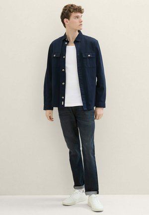 Marineblaues Hemdjackett mit zwei Brusttaschen, silbernen Knöpfen und einer lässigen Passform, kombiniert mit dunklen Jeans und weißen Sneakern.