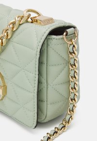 Pikowana torba na ramię w miętowym zielonym kolorze, ze złotą łańcuchem i okuciami, z płaskim złotym emblematem na przodzie. Kompaktowy prostokątny kształt.