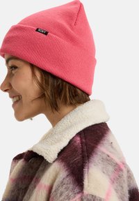 Roze geribde gebreide muts met een puntige bovenkant, voorzien van een klein zwart "ROXY" label; gedragen met een crèmekleurige en paarse geblokte jas.