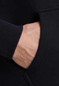 Zwarte sweatshirtstof met geribbelde boorden, met een zak aan de voorkant. Hand gedeeltelijk zichtbaar die in de zak rust.