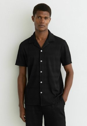 Jonge man draagt een zwart textuur kortärmelig overhemd met knopen en bijpassende broek, staat met een hand in de zak tegen een witte muur.