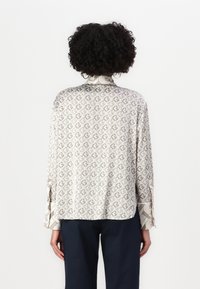 Long-sleeve blouse in light beige with a repeating geometric pattern, featuring a collar and contrasting patterned cuffs. Smooth fabric texture.