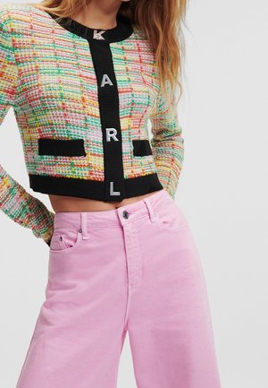 Femme portant un cardigan court tissé multicolore avec des bordures noires et des lettres argentées KARL, associé à un pantalon taille haute rose clair.