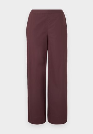 Jeune femme aux cheveux noirs et lisses portant une chemise bordeaux oversize, un pantalon large assorti, des lunettes et des chaussures noires, debout sur un sol gris.
