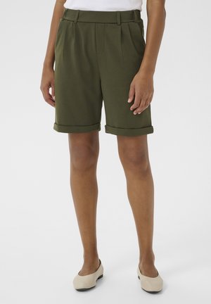 Persona che indossa pantaloncini verde oliva al ginocchio con risvolti arrotolati e scarpe basse beige, in piedi davanti a uno sfondo semplice.