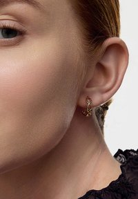 Złote kolczyki z plecionym wzorem pętli i małymi, bursztynowymi koralikami. Na neutralnym tle, które podkreśla ich detale.