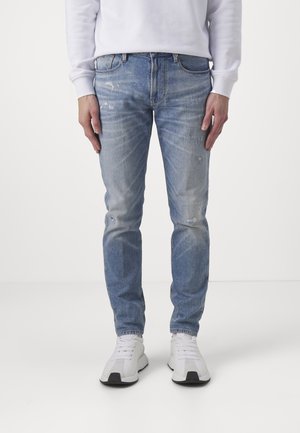 Person trägt hellblaue Slim-Fit-Jeans mit leichter Used-Optik, ein weißes Langarmshirt und weiße Sneaker, steht vor einfachem Hintergrund.