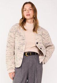 Veste beige oversized en mélange de laine avec un motif texturé, trois boutons et deux poches avant, portée sur un pull léger et un pantalon gris.