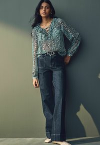 Blouse légère à motifs teal avec des volants, associée à un jean flare en denim foncé taille haute. Proximité d'un mur vert texturé.