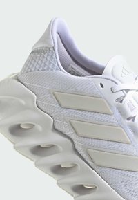 Białe buty sportowe z teksturowanym siateczkowym wierzchem, wyściełanym kołnierzem oraz charakterystyczną podeszwą z okrągłymi wycięciami i trzema poziomymi paskami.