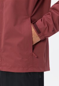 Giacca leggera color marrone scuro con finitura testurizzata, tasca laterale e polsini regolabili con dettagli in velcro, abbinata a pantaloni neri.