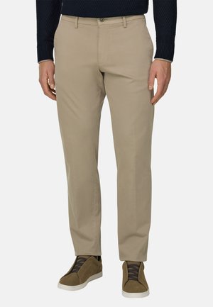 Homme portant un chino beige, des baskets vert olive avec des lacets noirs, et un pull texturé bleu foncé, debout devant un fond uni.
