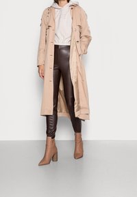 Beige trenčkot s knoflíky, v kombinaci s lesklými hnědými legínami a béžovými lodičkami se špičatou špičkou. Mikina s kapucí pod ním.