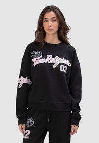 Czarna, krótka bluza z dużymi różowymi napisami "True Religion" i numerem "02" na przodzie oraz okrągłą naszywką z logo na lewym rękawie.