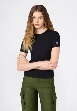 Młoda kobieta z długimi, falującymi włosami, ubrana w czarną koszulkę z krótkim rękawem i zielone spodnie typu cargo, ramiona skrzyżowane, neutralny wyraz twarzy, białe tło.