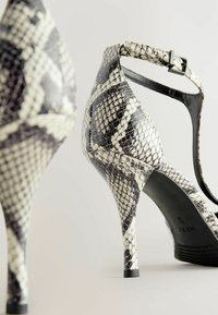 Juodos ir baltos python rašto aukštakulnės basutės su lieknu stiletto kulnu, turinčios kulkšnies dirželį ir tekstūruotą paviršių.