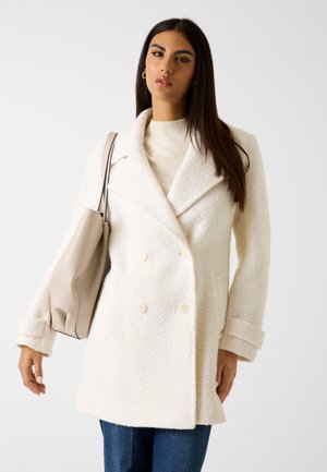 Manteau en laine blanc à double boutonnage avec une finition texturée, grands revers et accents de boutons. Associé à un sac à main beige clair.