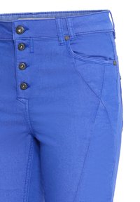 Gros plan d’un pantalon bleu avec quatre boutons visibles, une poche avant, des passants de ceinture et des coutures détaillées en haut à droite.