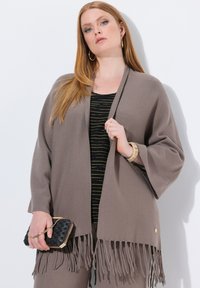 Chaqueta de punto marrón con flecos, hecha de un tejido suave, diseño abierto en la parte delantera, llevada sobre una camiseta negra a rayas. Accesorios incluyen un bolso negro texturizado.