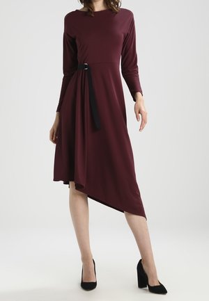 Nainen pukeutuneena pitkiin hihoihin, burgundinväriseen midi-mittaiseen mekkoon, jossa on epäsymmetrinen helma ja musta vyö, yhdistetty mustiin lohkokorkoihin.