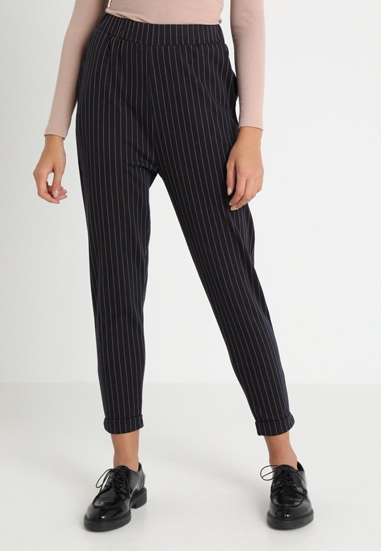 Femme portant un pantalon noir à fines rayures, fuselé avec ourlets retournés et chaussures noires à lacets, debout devant un fond blanc.