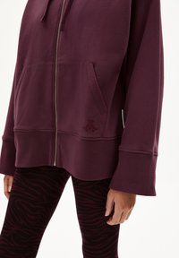 Bordeaux zip-up hoodie met een voorzak, voorzien van een geborduurd logo. Gepaard met zwarte legging met een paars zebra-strepenpatroon.