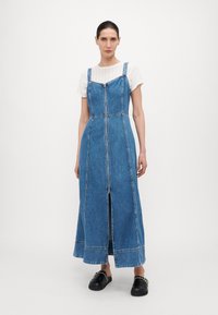 Denimmekko, jossa on kapeat olkaimet, edessä vetoketju ja sivuhalkio. Vaalea sininen väri ja kontrastiväriset saumat. Päällä valkoinen paita. Mustat liukuvat kengät.