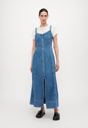 Denimkjole med smale stropper, glidelås foran og splitt i siden. Lys blå farge og kontrastsying. Båret over en hvit topp. Svart slipsko.