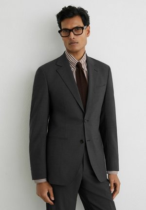 Mees, kes kannab tumedat halli ülikonda, pruunide triipudega särk, tumepruune lipsu ja musti ristkülikukujulisi prille, seisab valge seina ääres.