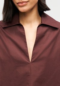 Chemise bordeaux avec col pointu, décolleté en V profond et texture lisse, avec une couture centrale décorative courant le long de l'avant.