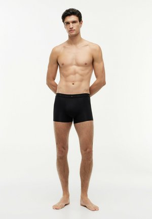 Szczupły męski model stoi boso, ubrany w czarne bokserki, ręce za plecami, neutralny wyraz twarzy, jednolite białe tło.